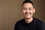 Professional headshot of a young Asian man in a black shirt, smiling warmly against a brown background